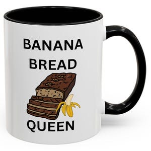 Könnte beinhalten: Weiße Keramiktasse mit schwarzem Rand und Henkel. Die Tasse trägt den Aufdruck "BANANA BREAD QUEEN" in Schwarz. Unter dem Text befindet sich ein Cartoon-Bild eines Bananenbrotlaibs und einer Bananenscheibe.