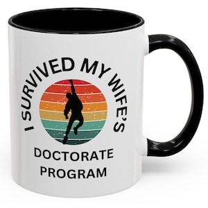 May include: White ceramic coffee mug with a black rim and handle. The mug has a retro-style graphic with the text "I survived my wife's doctorate program".