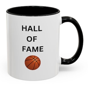 May include: White ceramic mug with a black interior and handle. The mug features the words "HALL OF FAME" in black block letters, along with a basketball graphic. A sports-themed gift.