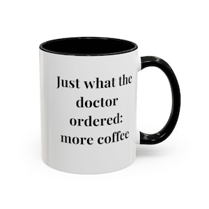May include: White ceramic coffee mug with a black rim and handle. The mug has black text that reads "Just what the doctor ordered: more coffee".