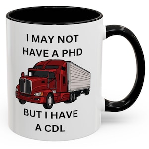Op de afbeelding: Een witte keramische koffiemok met een zwarte rand en handvat. De mok heeft een rode vrachtwagen met een witte aanhanger erop. De tekst op de mok luidt "I may not have a PHD but I have a CDL".