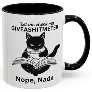 Op de afbeelding: Een witte keramische koffiemok met een zwarte rand en handvat. De mok heeft een zwart-witte illustratie van een kat die met een boek en een kopje koffie zit. De tekst op de mok is "Let me check my GIVEASHITMETER Nope, Nada".