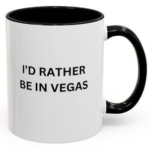 May include: White ceramic coffee mug with a black rim and handle. The mug has the text "I'd rather be in Vegas" printed in black.