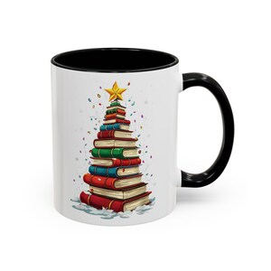May include: White ceramic mug with a black interior and handle. The mug features a Christmas tree design made of stacked books in various colors, topped with a gold star. Colorful confetti surrounds the tree.