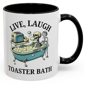 May include: White ceramic coffee mug with a black rim and handle. The mug features a black and white illustration of a skeleton taking a bath in a blue bathtub with bubbles. The text "Live, Laugh, Toaster Bath" is printed on the mug.