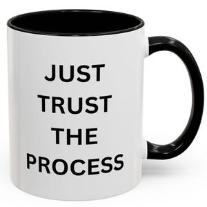 Op de afbeelding: Witte keramische koffiemok met een zwarte rand en handvat. De mok heeft zwarte tekst met de tekst "JUST TRUST THE PROCESS".
