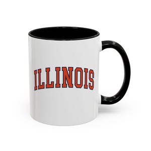 May include: White ceramic mug with a black interior and handle. The word "ILLINOIS" is printed in orange block letters with a blue outline, centered on the mug.