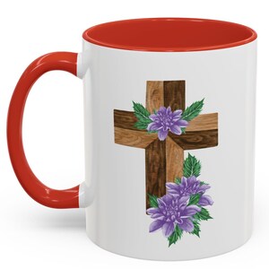Puede incluir: Taza de cerámica blanca con interior y asa rojos. La taza presenta un diseño de cruz de madera adornada con flores moradas y hojas verdes. El diseño está centrado en el exterior de la taza.