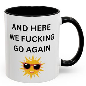 Pode incluir: Caneca de cerâmica branca com interior e alça pretos. A caneca apresenta o texto "AND HERE WE FUCKING GO AGAIN" em preto, com um sol sorrindo usando óculos de sol abaixo do texto. Um design humorístico e relacionável.