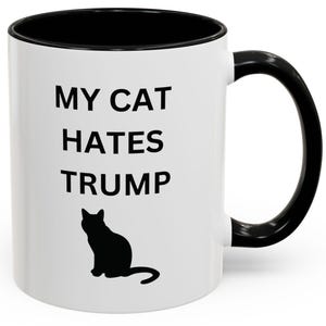 May include: White ceramic mug with a black interior and handle. The mug features the text "MY CAT HATES TRUMP" in black, along with a black silhouette of a cat.