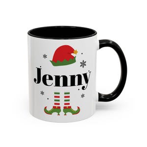 May include: White ceramic mug with a black interior and handle. The mug features the name "Jenny" in black script, along with a red and green elf hat, snowflakes, and elf feet. A festive holiday-themed design.
