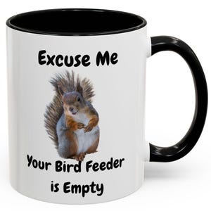 May include: White ceramic coffee mug with a black rim and handle. The mug features a squirrel with the text "Excuse Me Your Bird Feeder is Empty".