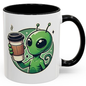 Puede incluir: Taza de café de cerámica blanca con borde y asa negros. La taza presenta una ilustración de dibujos animados de un extraterrestre verde sosteniendo una taza de café para llevar.