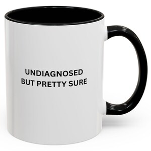 Könnte beinhalten: Weiße Keramik-Kaffeetasse mit schwarzem Rand und Henkel. Die Tasse hat einen schwarzen Schriftzug mit der Aufschrift "Undiagnosed but pretty sure".