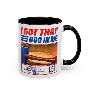 May include: White ceramic mug with a black interior and handle. The mug features the text "I GOT THAT DOG IN ME" in red and blue. Below the text is an image of a hot dog and soda. The mug also has the text "¼ POUND PLUS ALL BEEF HOT DOG AND 20 oz. SODA (With Refill) 580 - 850 cal."