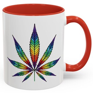 Puede incluir: Una taza de café de cerámica blanca con un borde y un asa rojos. La taza presenta un diseño de hoja de marihuana arcoíris colorido.