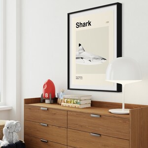 May include: A framed print featuring a shark illustration with the word "Shark" at the top. The print is displayed above a wooden dresser with a white lamp, a red rocket, and books. A wicker basket and stuffed animal are in the foreground.