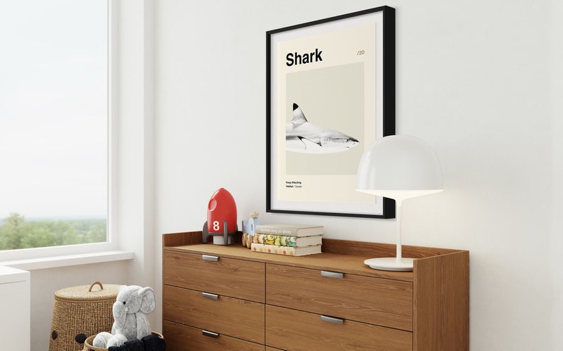 May include: A framed print featuring a shark illustration with the word "Shark" at the top. The print is displayed above a wooden dresser with a white lamp, a red rocket, and books. A wicker basket and stuffed animal are in the foreground.