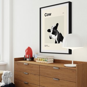 May include: A black framed poster with a white background and a black and white image of a cow's head. The poster has the word "Cow" in black text at the top and the number "178" in smaller black text to the right of the word "Cow". The poster is hanging on a white wall above a wooden dresser with six drawers.