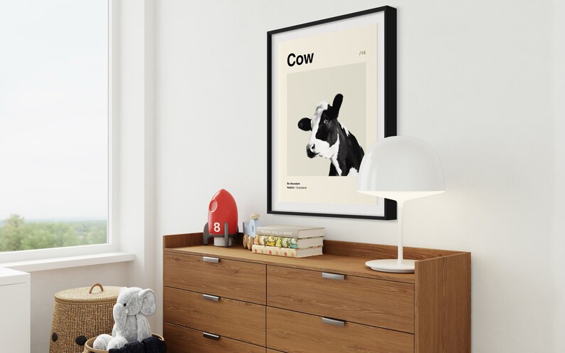 May include: A black framed poster with a white background and a black and white image of a cow's head. The poster has the word "Cow" in black text at the top and the number "178" in smaller black text to the right of the word "Cow". The poster is hanging on a white wall above a wooden dresser with six drawers.