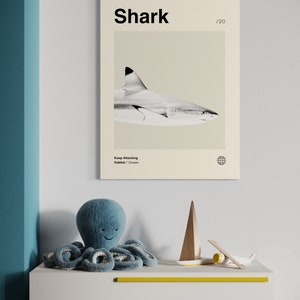 May include: A framed print featuring a grayscale image of a shark with the word "Shark" at the top. Below the print is a blue plush octopus toy, a wooden sailboat, and a small airplane on a white dresser with yellow accents.