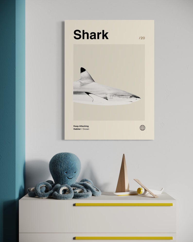 May include: A framed print featuring a grayscale image of a shark with the word "Shark" at the top. Below the print is a blue plush octopus toy, a wooden sailboat, and a small airplane on a white dresser with yellow accents.