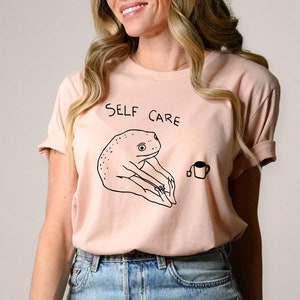 May include: A light pink t-shirt with a black and white illustration of a frog sitting with a cup of tea. The text "SELF CARE" is printed on the shirt.