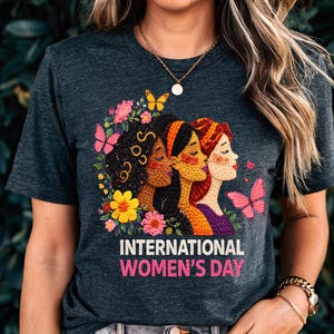 May include: Dark gray t-shirt featuring a colorful graphic of three women's faces surrounded by flowers and butterflies. The text "INTERNATIONAL WOMEN'S DAY" is printed in pink below the graphic. The shirt has a crew neck.