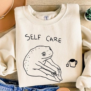 May include: A cream-colored sweatshirt with the text "SELF CARE" and a black line drawing of a frog holding a cup of tea.