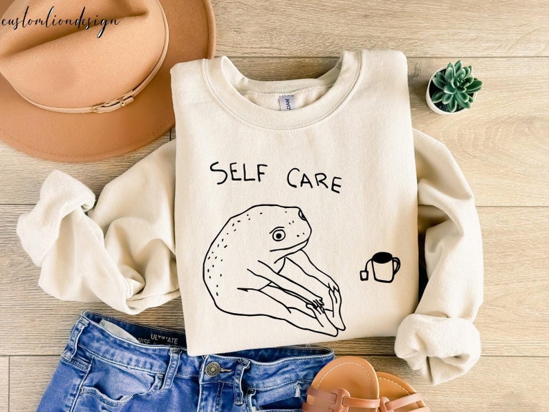 May include: A cream-colored sweatshirt with the text "SELF CARE" and a black line drawing of a frog holding a cup of tea.