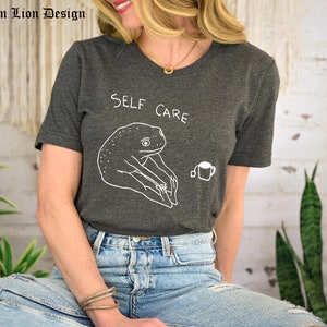 May include: A dark gray t-shirt with the text "SELF CARE" and a cartoon frog drinking tea.