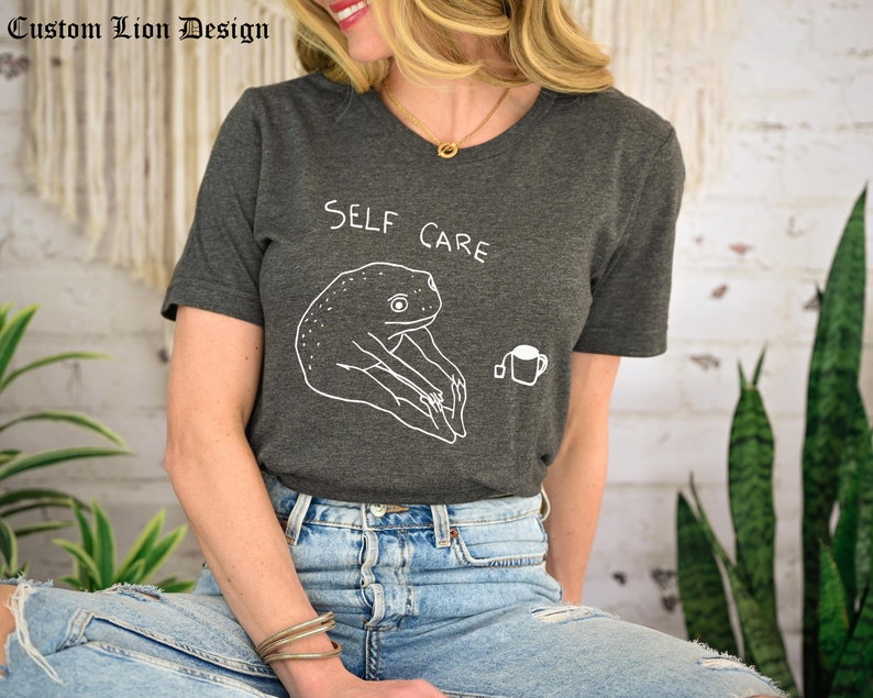 May include: A dark gray t-shirt with the text "SELF CARE" and a cartoon frog drinking tea.