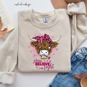 K&ouml;nnte beinhalten: Ein beiger Pullover mit einer rosa und schwarzen Highland-Kuh, die ein rosa Band und ein Bandana tr&auml;gt. Der Text "pray believe fight" ist in rosa unter der Kuh aufgedruckt.