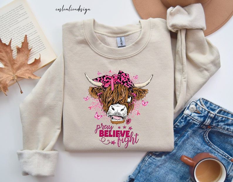 K&ouml;nnte beinhalten: Ein beiger Pullover mit einer rosa und schwarzen Highland-Kuh, die ein rosa Band und ein Bandana tr&auml;gt. Der Text "pray believe fight" ist in rosa unter der Kuh aufgedruckt.