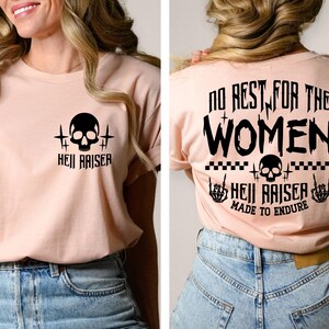 May include: A light pink t-shirt featuring a black graphic on the front left chest of a skull and the text "HELL RAISER". The back displays the text "NO REST FOR THE WOMEN", a skull, checkered patterns, and "HELL RAISER MADE TO ENDURE".