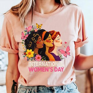 May include: A light pink t-shirt featuring a colorful embroidered design for International Women's Day. The design includes three women's faces, flowers, and butterflies. The text "INTERNATIONAL WOMEN'S DAY" is printed below the design.