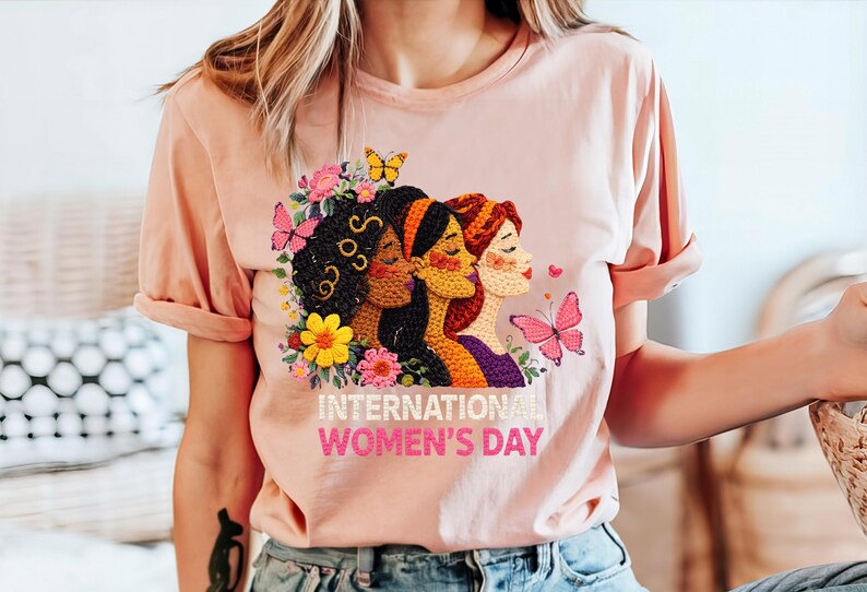 May include: A light pink t-shirt featuring a colorful embroidered design for International Women's Day. The design includes three women's faces, flowers, and butterflies. The text "INTERNATIONAL WOMEN'S DAY" is printed below the design.