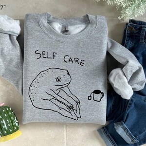 May include: Gray sweatshirt with a black frog illustration and the text "SELF CARE" printed on the front. A small black cup with a tea bag is also illustrated on the sweatshirt.