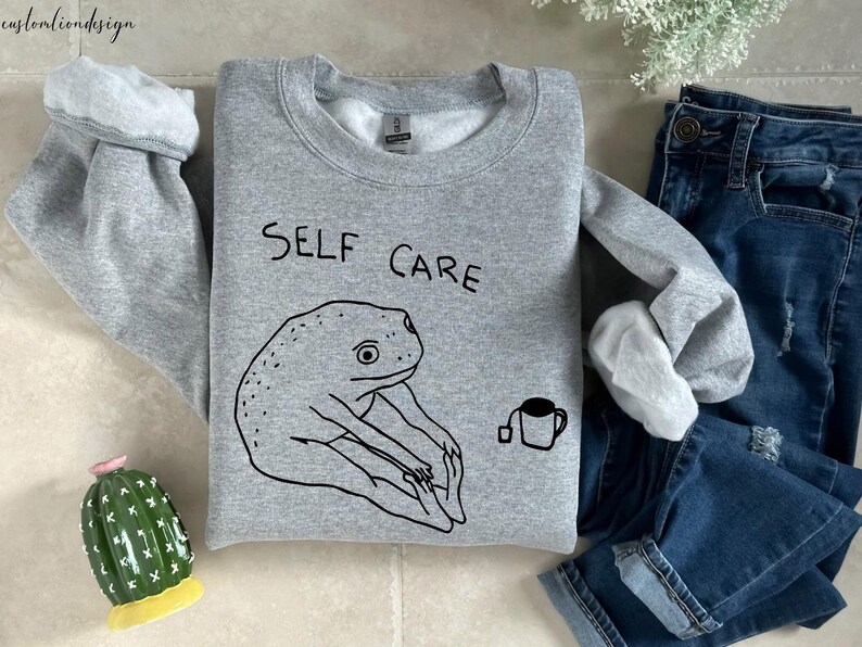 May include: Gray sweatshirt with a black frog illustration and the text "SELF CARE" printed on the front. A small black cup with a tea bag is also illustrated on the sweatshirt.