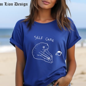 May include: A royal blue t-shirt with a white outline of a frog holding a cup of tea and the text "SELF CARE".