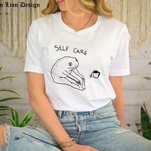 May include: White t-shirt with a black line drawing of a frog doing yoga and the text "SELF CARE". A small cup of tea is drawn next to the frog.