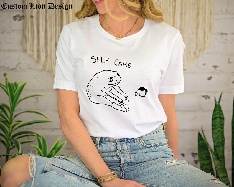 May include: White t-shirt with a black line drawing of a frog doing yoga and the text "SELF CARE". A small cup of tea is drawn next to the frog.