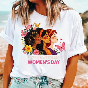 May include: White t-shirt featuring a colorful graphic celebrating International Women's Day. The design includes three women's faces, butterflies, and floral elements. The text "INTERNATIONAL WOMEN'S DAY" is displayed in pink.