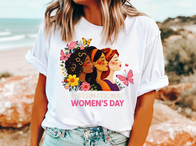 May include: White t-shirt featuring a colorful graphic celebrating International Women's Day. The design includes three women's faces, butterflies, and floral elements. The text "INTERNATIONAL WOMEN'S DAY" is displayed in pink.
