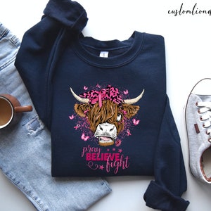 K&ouml;nnte beinhalten: Ein marineblaues Sweatshirt mit einem rosa Band und einer Highland-Kuh-Grafik. Der Text "beten glauben k&auml;mpfen" ist unter der Kuh gedruckt.
