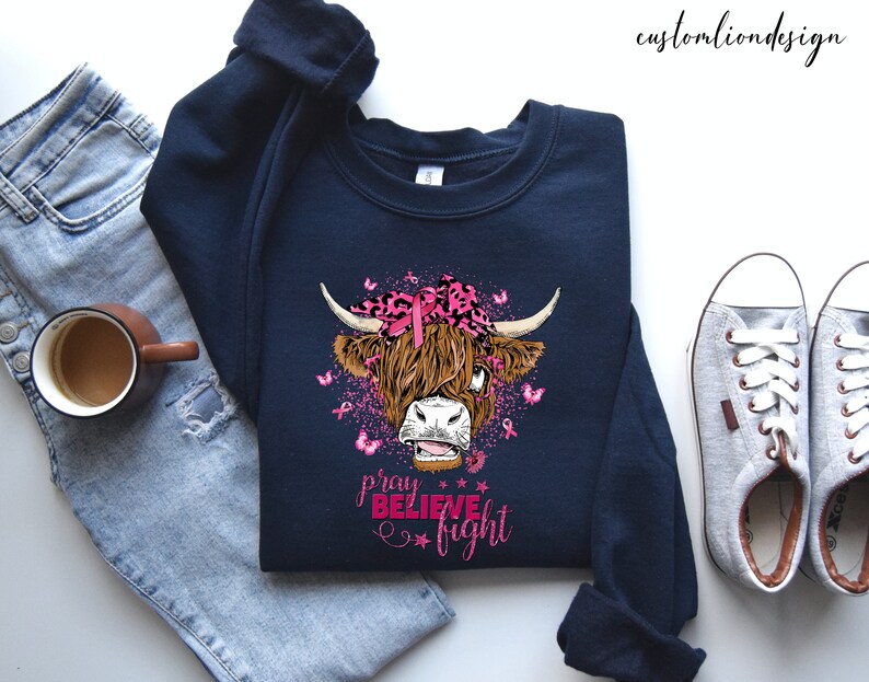 K&ouml;nnte beinhalten: Ein marineblaues Sweatshirt mit einem rosa Band und einer Highland-Kuh-Grafik. Der Text "beten glauben k&auml;mpfen" ist unter der Kuh gedruckt.