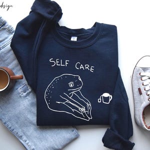 May include: A dark blue sweatshirt with a white frog graphic and the text "SELF CARE". The frog is holding a cup of tea.