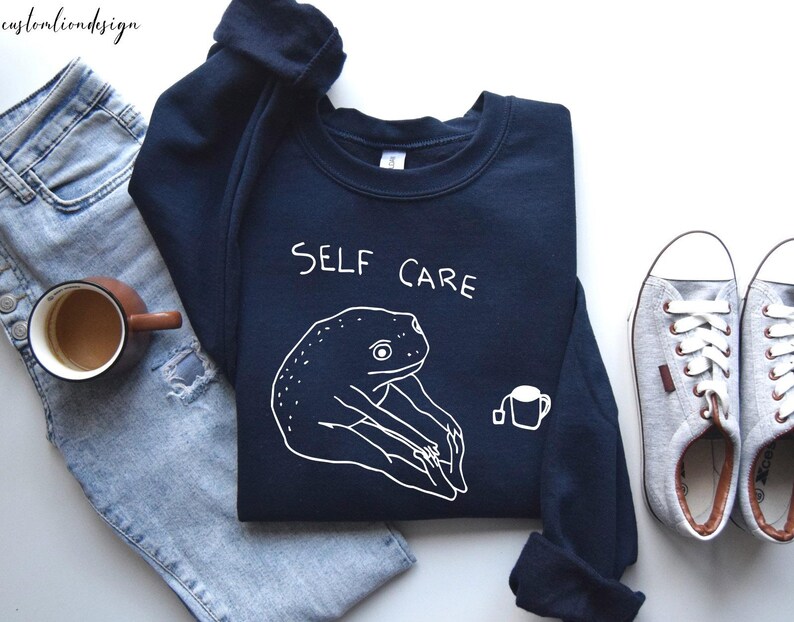 May include: A dark blue sweatshirt with a white frog graphic and the text "SELF CARE". The frog is holding a cup of tea.