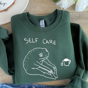 May include: A dark green sweatshirt with the text "SELF CARE" in white lettering. A white outline drawing of a frog sitting in a yoga pose with a cup of tea is below the text.