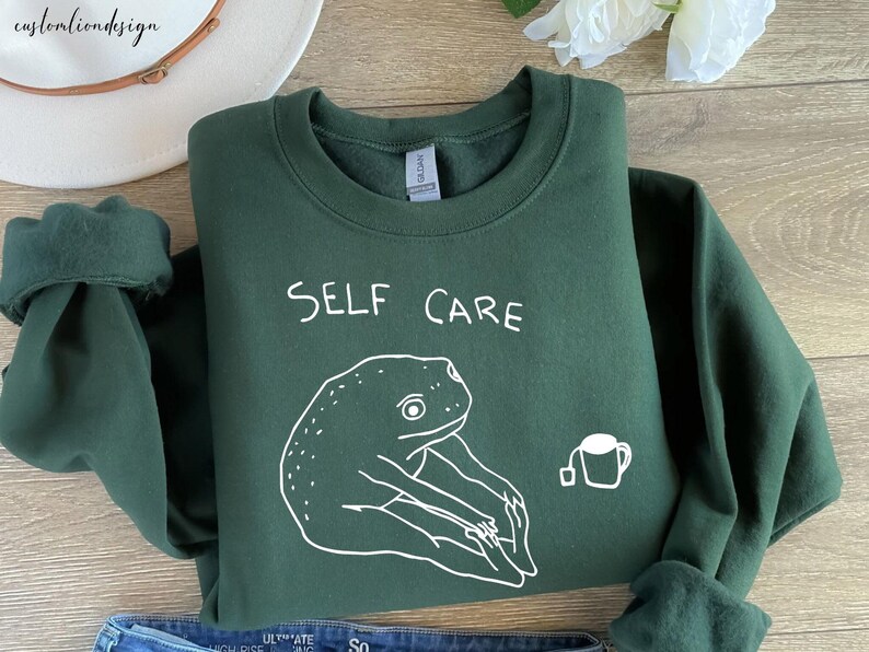 May include: A dark green sweatshirt with the text "SELF CARE" in white lettering. A white outline drawing of a frog sitting in a yoga pose with a cup of tea is below the text.